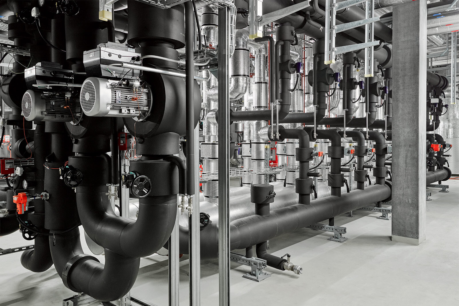 Pumpen mit Motoren und rot markierten Armaturen an schwarz gedämmten Rohrleitungen