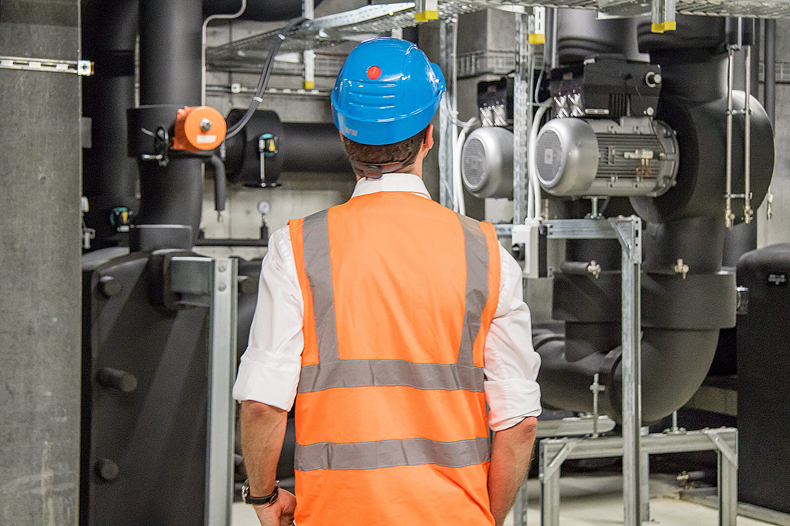 Fachperson in Warnweste und Helm, von hinten gesehen, vor Pumpen und Rohrleitungen einer Technikzentrale
