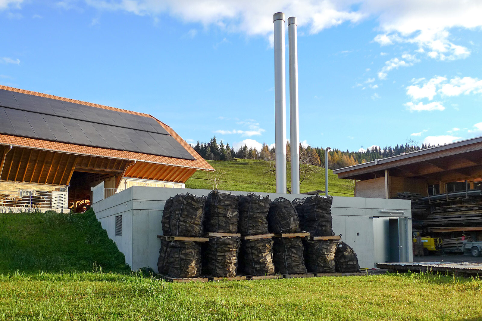 Heizzentrale des Nahwärmeverbunds Zugerberg mit zwei Kaminen, davor Holzvorrat, daneben ein Scheunendach mit Solarpanels